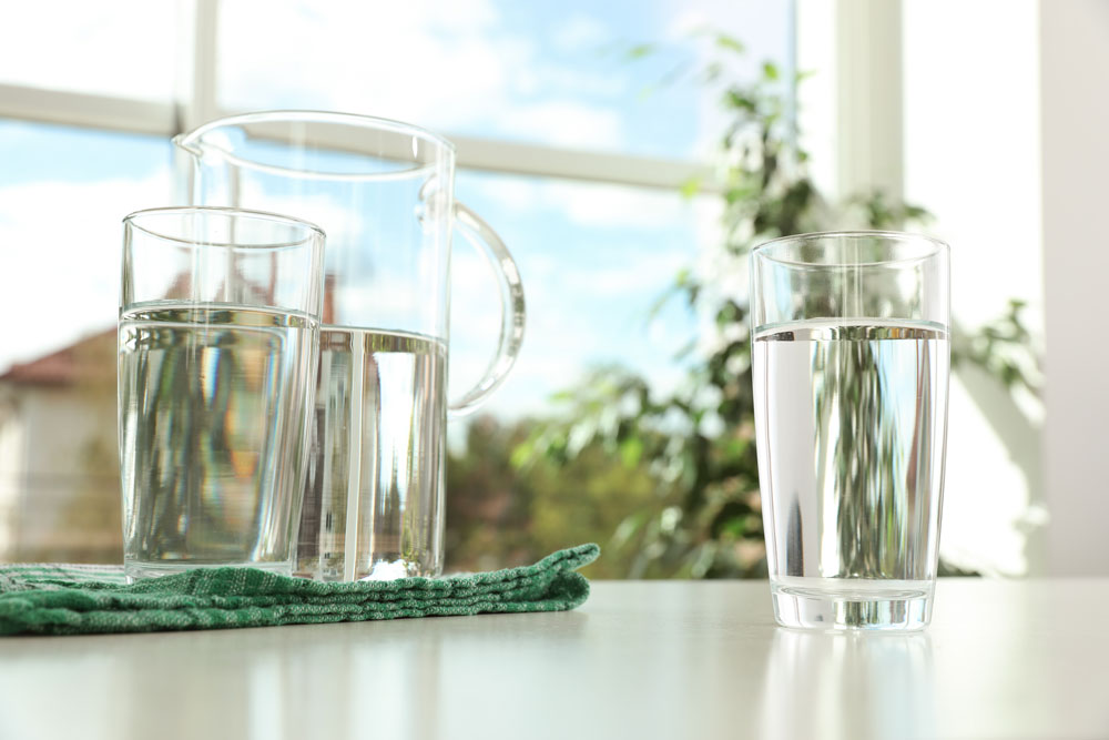 Full water pitcher and glasses on a table in front of a window